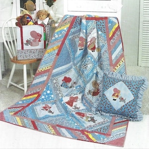 Puede incluir: Una colcha de retazos azul y roja con un borde blanco. La colcha presenta un patrón repetitivo de niñas pequeñas con vestidos rojos y gorros blancos. La colcha se exhibe en una cómoda blanca con una almohada en el suelo.