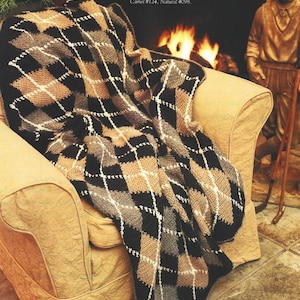 May include: A black, gray, and brown plaid crocheted throw blanket draped over a tan armchair. The blanket is made with alpaca yarn. The text "Project was stitched with Al-pa-ka: Black #153, Oxford Grey #152, Camel #124, Natural #098." is visible in the image.