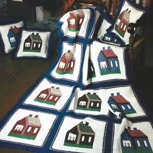May include: A crocheted blanket with a white background and blue borders features a repeating pattern of colorful houses. The blanket is folded over a wooden chair with two smaller crocheted pillows with the same house pattern.