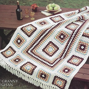 May include: Off-white crochet granny square afghan with brown, yellow and blue accents. The afghan is draped over a wooden picnic table.