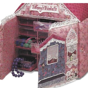 K&ouml;nnte beinhalten: Eine rosa Stoff-Schmuckschatulle in Form eines Hauses mit einer wei&szlig;en Spitzenborte und den aufgestickten Worten "Tiffany's Trinkets" auf der Vorderseite. Die Schatulle hat mehrere Schubladen zum Verstauen von Kleinigkeiten.