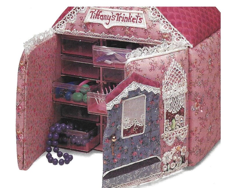 K&ouml;nnte beinhalten: Eine rosa Stoff-Schmuckschatulle in Form eines Hauses mit einer wei&szlig;en Spitzenborte und den aufgestickten Worten "Tiffany's Trinkets" auf der Vorderseite. Die Schatulle hat mehrere Schubladen zum Verstauen von Kleinigkeiten.