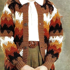 May include: A crochet cardigan in shades of brown, white, and orange. The long-sleeved garment features a button-up front and a textured pattern. The person is wearing a white collared shirt and a brown belt.