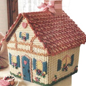 May include: A crocheted house-shaped tissue box cover with a pink roof, blue door, and yellow windows. The house is decorated with flowers and hearts.