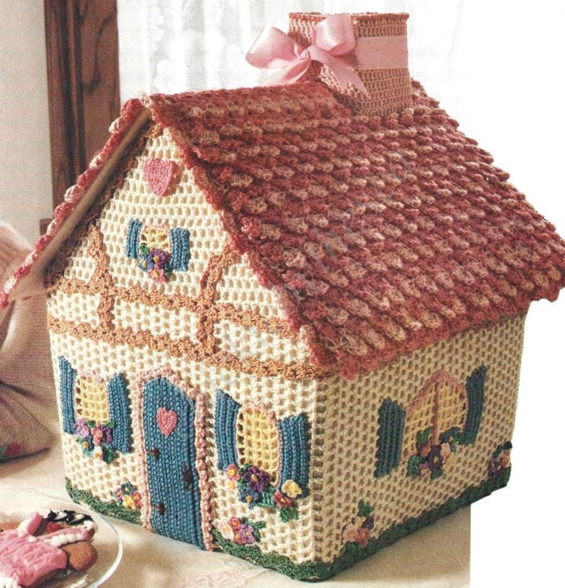 May include: A crocheted house-shaped tissue box cover with a pink roof, blue door, and yellow windows. The house is decorated with flowers and hearts.