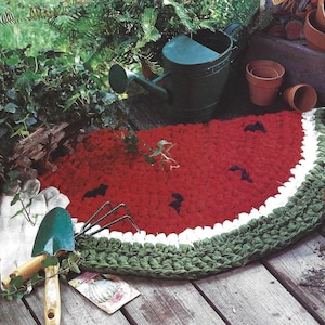 May include: A red, white, and green crocheted watermelon rug with black seed accents. The rug is on a wooden deck with gardening tools and potted plants.