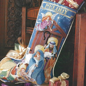 May include: A blue and gold Christmas stocking featuring a nativity scene and the word "MELODY" embroidered at the top. The stocking is filled with gold ribbon and a small gift box.