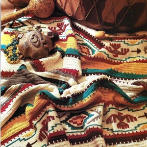 May include: A vibrant, hand-crocheted blanket with a Southwestern pattern. The blanket displays geometric designs in cream, brown, red, yellow, and teal. A wooden drum, maracas, and other decorative items are arranged on top of the blanket.