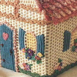 May include: A crocheted house-shaped basket with a pink roof, blue door, and yellow windows. The basket is decorated with crocheted flowers and green leaves.