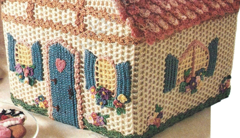 May include: A crocheted house-shaped basket with a pink roof, blue door, and yellow windows. The basket is decorated with crocheted flowers and green leaves.