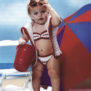 May include: A young child wearing a red and white striped crochet swimsuit and a white crochet cardigan. The child is holding a red bucket and wearing sunglasses. The child is standing in front of a white beach chair and a red and blue beach umbrella.