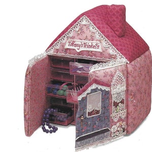 K&ouml;nnte beinhalten: Eine rosa und florale Stoff-Schmuckbox in Hausform mit einer Klappt&uuml;r und mehreren Schubladen. Die Box ist mit Spitze verziert und die Worte "Tiffany's Trinkets" sind auf der Vorderseite gestickt.