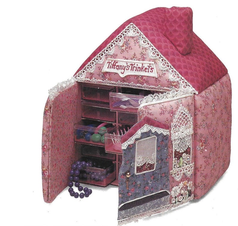 K&ouml;nnte beinhalten: Eine rosa und florale Stoff-Schmuckbox in Hausform mit einer Klappt&uuml;r und mehreren Schubladen. Die Box ist mit Spitze verziert und die Worte "Tiffany's Trinkets" sind auf der Vorderseite gestickt.