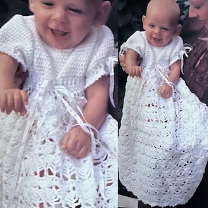 Könnte beinhalten: Weißes, handgefertigtes Babykleid aus Häkelarbeit mit kurzen Ärmeln und einem fließenden Rock. Das Kleid hat komplizierte Lochmuster und ist mit Satinbändern an den Ärmeln und in der Taille verziert. Das Baby lächelt.