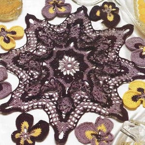 May include: A handmade crochet doily in dark purple with a starburst design. Surrounding the doily are several crochet pansies in purple and yellow. The doily and flowers are placed on a white satin surface.