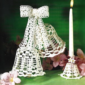 May include: A decorative arrangement featuring two white crocheted bell ornaments and a lit candle. The bells are suspended from a bow and adorned with delicate lace patterns. The candle is placed in a matching crocheted holder, with floral accents.