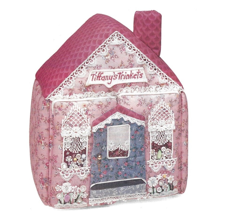 K&ouml;nnte beinhalten: Eine rosa und blaue Stoff-Schmuckdose in Form eines Hauses mit Spitzenbesatz und dem Schriftzug "Tiffany's Trinkets" auf der Vorderseite.