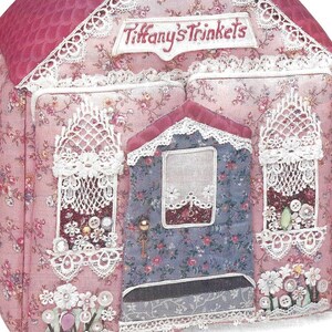 K&ouml;nnte beinhalten: Ein rosa Stoffhaus mit wei&szlig;em Spitzenbesatz und dem Schriftzug "Tiffany's Trinkets", der auf die Vorderseite gestickt ist. Das Haus hat drei Fenster mit wei&szlig;en Spitzenvorh&auml;ngen und eine T&uuml;r mit einem Schl&uuml;sselloch. Das Haus ist mit Perlen, Kn&ouml;pfen und Blumen verziert.