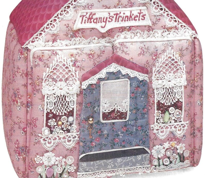 K&ouml;nnte beinhalten: Ein rosa Stoffhaus mit wei&szlig;em Spitzenbesatz und dem Schriftzug "Tiffany's Trinkets", der auf die Vorderseite gestickt ist. Das Haus hat drei Fenster mit wei&szlig;en Spitzenvorh&auml;ngen und eine T&uuml;r mit einem Schl&uuml;sselloch. Das Haus ist mit Perlen, Kn&ouml;pfen und Blumen verziert.