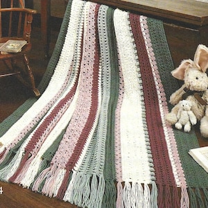 Puede incluir: Una manta de punto de rayas con flecos en tonos verdes, blancos, rosas y burdeos. La manta está drapeada sobre una superficie de madera y un conejito de peluche está sentado sobre la manta.