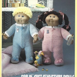 May include: Two soft sculpture dolls, one in blue and one in pink, wearing crocheted pyjamas. The dolls have yarn hair and are holding hands. The text at the bottom reads "FOR 16" SOFT SCULPTURE DOLLS."