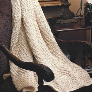 May include: A cream-colored crocheted throw blanket draped over a wooden armchair. The blanket has a textured, cable knit pattern.