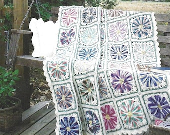 Op de afbeelding: Een gehaakt deken met een witte achtergrond en kleurrijke bloemmotieven. Het deken is gemaakt van garen in tinten blauw, paars, roze, groen en bruin. Het deken is gevouwen en gedrapeerd over een houten bank.