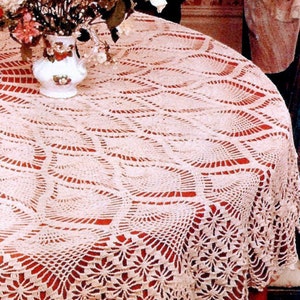 May include: A white crocheted tablecloth with a delicate, intricate pattern. The tablecloth is laid over a red table with a floral arrangement in a white vase.