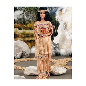 Peut inclure: Une poupée amérindienne portant une robe en crochet avec des franges et des accents de perles. La poupée a de longs cheveux noirs et porte un bandeau avec une plume. La poupée est debout sur une surface brune avec des rochers et un fond de fourrure blanche.