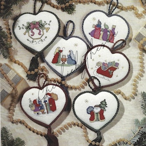 May include: Five white heart-shaped Christmas decorations with cross-stitch designs. The decorations feature a nativity scene, Santa Claus, a sleigh with presents, a bell, and a Christmas tree. Each decoration has a black tassel.