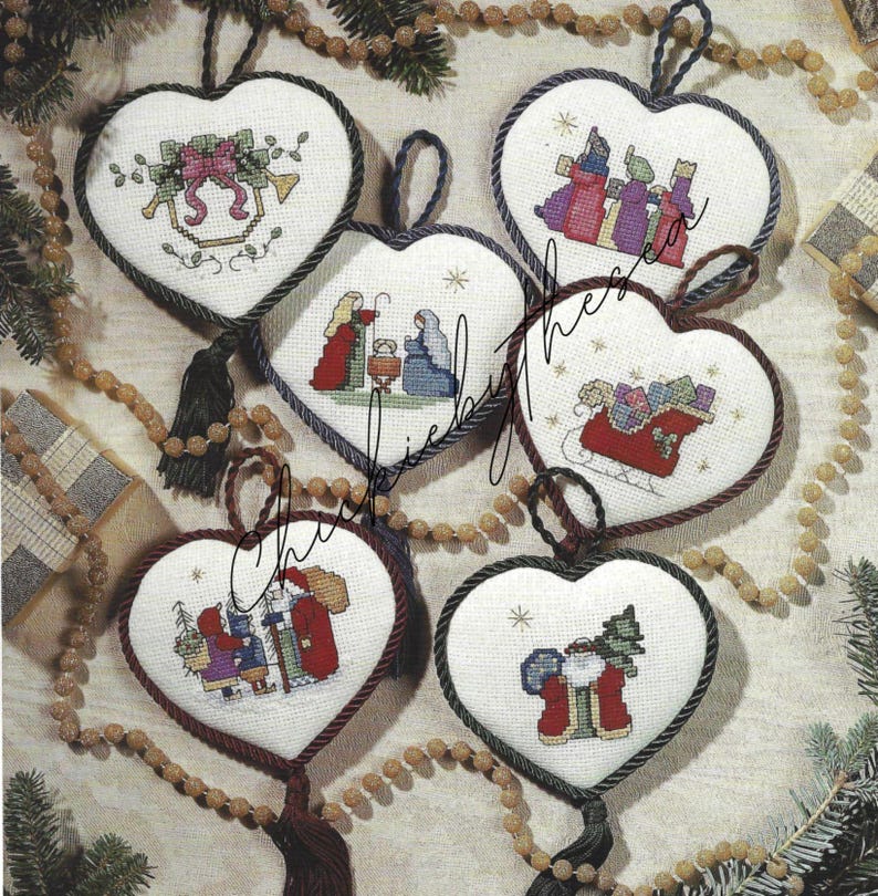 May include: Five white heart-shaped Christmas ornaments with cross-stitch designs. The ornaments feature a nativity scene, Santa Claus, a sleigh with presents, a bell, and a Christmas tree. Each ornament has a black tassel.