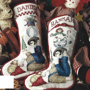 Può includere: Due calze di Natale personalizzate con bordo a quadri rossi e bianchi. La calza a sinistra ha il nome "DANIEL" ricamato e presenta un'illustrazione fantasiosa di un giullare che tiene in equilibrio una palla, una scatola di giocattoli e un albero di Natale. La calza a destra ha il nome "HANNAH" ricamato e presenta un'illustrazione fantasiosa di una bambola di pezza che tiene in equilibrio una palla, una scatola di giocattoli e una canna di zucchero.