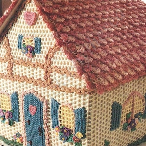 May include: A crocheted house shaped basket with a pink roof, blue door, and yellow windows. The house has a white base with brown trim and floral accents.