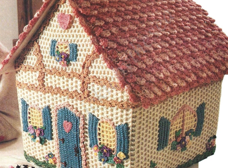 May include: A crocheted house shaped basket with a pink roof, blue door, and yellow windows. The house has a white base with brown trim and floral accents.