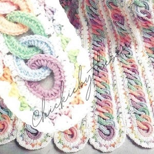 May include: A crocheted baby blanket with a white background and colorful stripes. The blanket features a unique design with interlocking loops in shades of pink, blue, green, and orange.