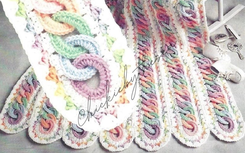 May include: A crocheted baby blanket with a white background and colorful stripes. The blanket features a unique design with interlocking loops in shades of pink, blue, green, and orange.