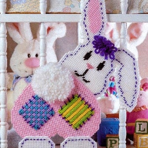 May include: A pink and white crocheted bunny with a purple flower on its head. The bunny has a white fluffy tail and is wearing a patchwork dress with green, yellow, blue, and purple squares.