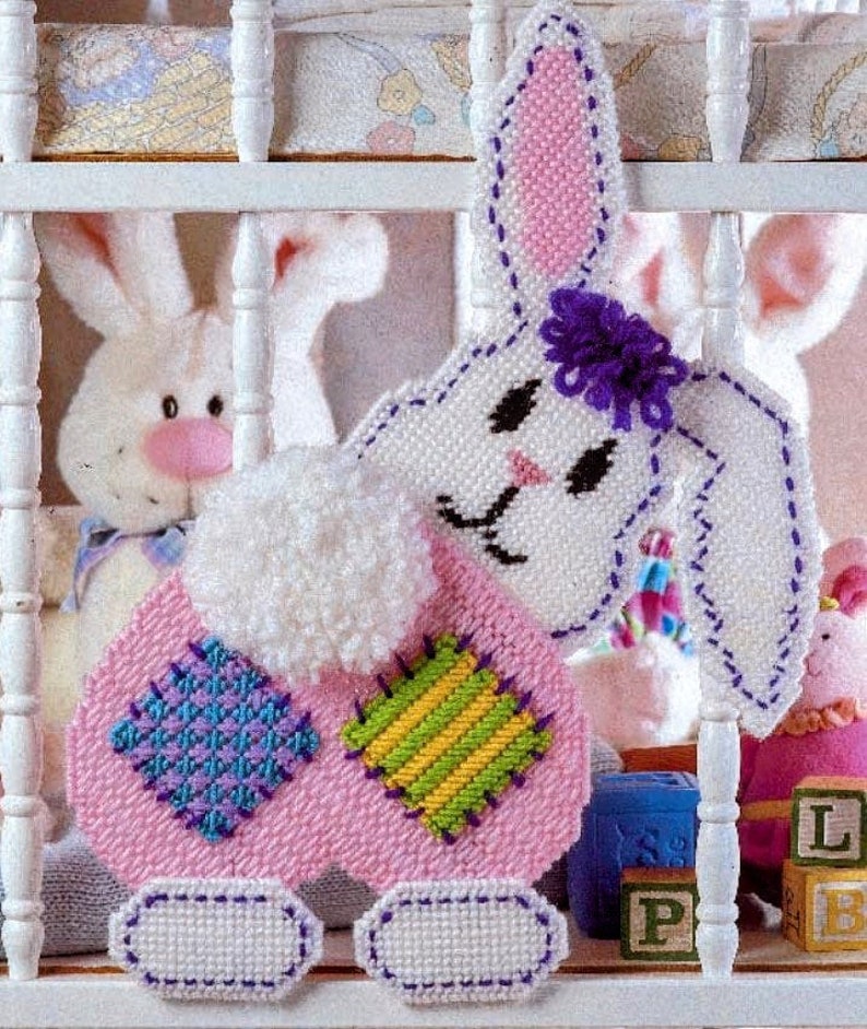 May include: A pink and white crocheted bunny with a purple flower on its head. The bunny has a white fluffy tail and is wearing a patchwork dress with green, yellow, blue, and purple squares.