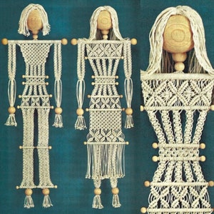 Pode incluir: Dois tapetes de macramé em forma de pessoas. As figuras são feitas com cordão branco e têm contas de madeira para a cabeça e os braços. As figuras estão penduradas em uma barra de madeira.