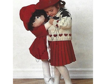 Könnte beinhalten: Ein junges Mädchen mit einem roten Hut und einem weißen Pullover mit roten Herzen hält eine rote Puppe mit einem roten Hut und einem roten Mantel. Das Mädchen trägt einen roten Rock und weiße Strumpfhosen. Die Puppe trägt ein weißes Kleid und weiße Strumpfhosen.