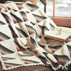 May include: A crocheted throw blanket with a diamond pattern in shades of green, red, and white. The blanket is draped over a brown leather armchair with a book and glasses on top.