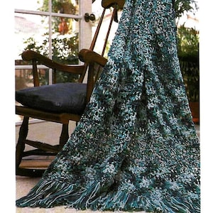 May include: A handmade, textured crochet blanket in shades of teal, green, and white, draped over a wooden rocking chair. The blanket features a fringed edge and a cozy, inviting appearance. The chair has a black cushion.