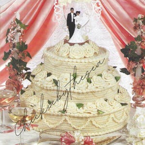 May include: A three-tiered, cream-coloured crocheted wedding cake adorned with cream roses and green leaves. A bride and groom cake topper sits on top. The cake is displayed with champagne glasses and floral arrangements.