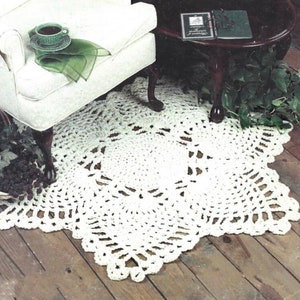 May include: A white crocheted square rug with a lacy design. The rug is placed on a wooden floor and is partially under a white armchair and a wooden table.