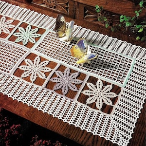 Puede incluir: Camino de mesa blanco de ganchillo con diseño floral. El camino de mesa presenta paneles cuadrados con motivos florales en tonos blanco, verde claro y lavanda. Dos figuras de mariposas están colocadas sobre el camino de mesa.