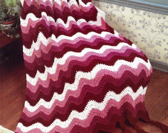 May include: A handmade crochet blanket in a chevron pattern. The blanket features alternating stripes of white, pink, and burgundy. The blanket is draped over a chair and has burgundy tassels along the bottom edge.