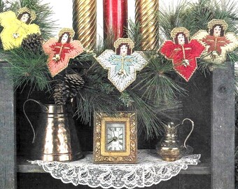 Può includere: Una ghirlanda di cinque ornamenti a forma di angelo ricamati a punto croce in giallo, pesca, azzurro, rosso e crema, esposti con una brocca metallica, un piccolo orologio rettangolare con cornice dorata e una lattiera argentata su un centrino di pizzo bianco.
