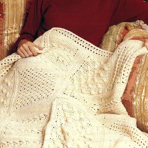 May include: Cream-colored, hand-crocheted blanket with a variety of textured stitch patterns. The blanket is draped over a person's lap, showcasing the intricate details of the yarn work. The blanket's design includes squares and geometric patterns.