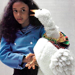 以下が含まれることがあります： 長い黒髪の若い女の子が、首にカラフルなフリルが付いた白いふわふわのガチョウの人形を持っています。人形は大きく開いた口をして、女の子を見ています。