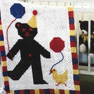 Op de afbeelding: Een wit gebreid babydekentje met een zwarte beer die een gele feestmuts draagt en rode en blauwe ballonnen vasthoudt. Een geel kuiken loopt rechts van de beer. Het dekentje heeft een rode, gele en blauwe rand.